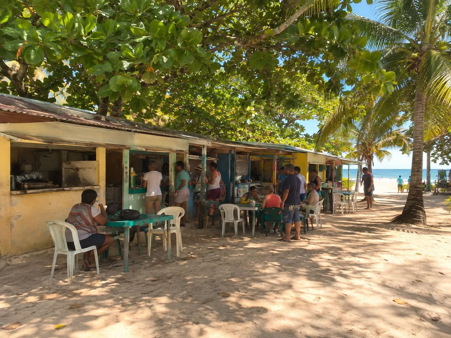 A row of faded plywood food kiosks under sea grape trees, plastic tables set out in front of them